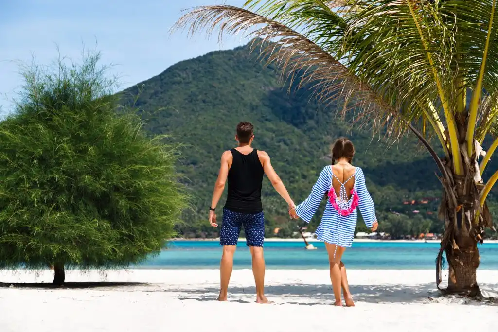 Couple holding hands on a white sand beach, standing barefoot near a palm tree with clear blue water and a lush green hillside in the background