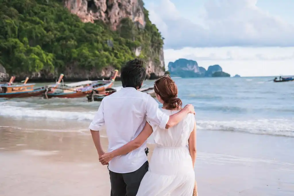 Couple walking arm-in-arm on a sandy beach, facing turquoise waters with traditional longtail boats and dramatic limestone cliffs in the background