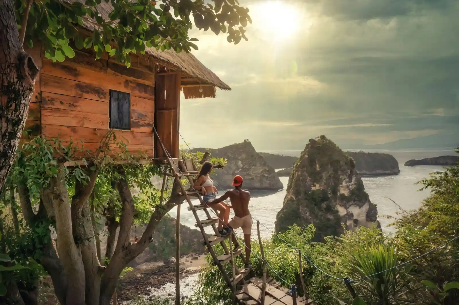 A couple stands on the wooden steps of a rustic cliffside treehouse, looking out at a dramatic ocean sunrise over rocky islands, highlighting romantic things to do for Valentine's Day in Bali.
