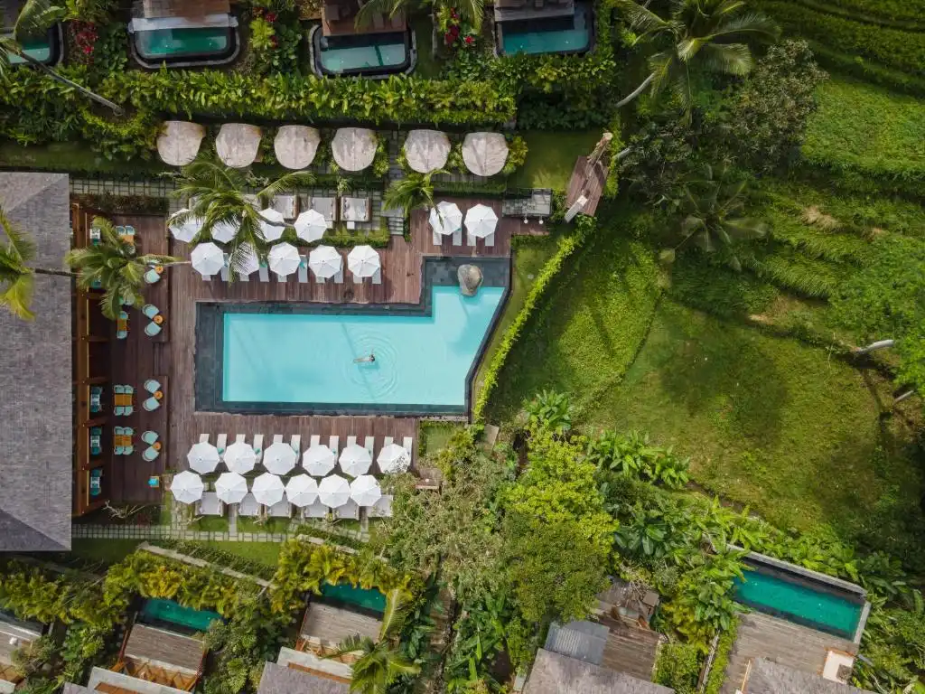 A top-down aerial view of a bright blue geometric swimming pool surrounded by a wooden sun deck and neat rows of white umbrellas, bordered by lush green terraces at one of the best luxury resorts in Bali.