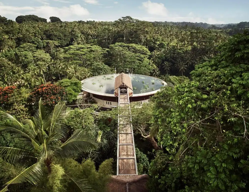 Drone photograph looking down at a long wooden bridge suspended high above tropical rainforest foliage, connecting to an elliptical reflecting pool at one of the best luxury resorts in Bali.