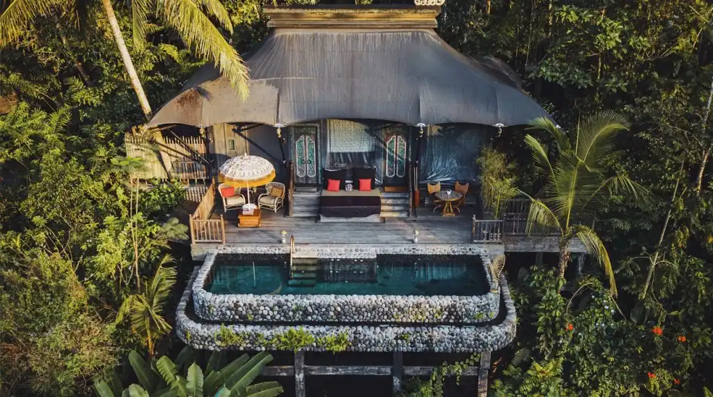 A luxurious jungle lodge with a large, draped canvas roof built among the trees. The villa features an outdoor lounging deck with a traditional Balinese umbrella, daybeds, and a private plunge pool made of smooth river stones, showcasing the unique architecture of the best luxury resorts in Bali.