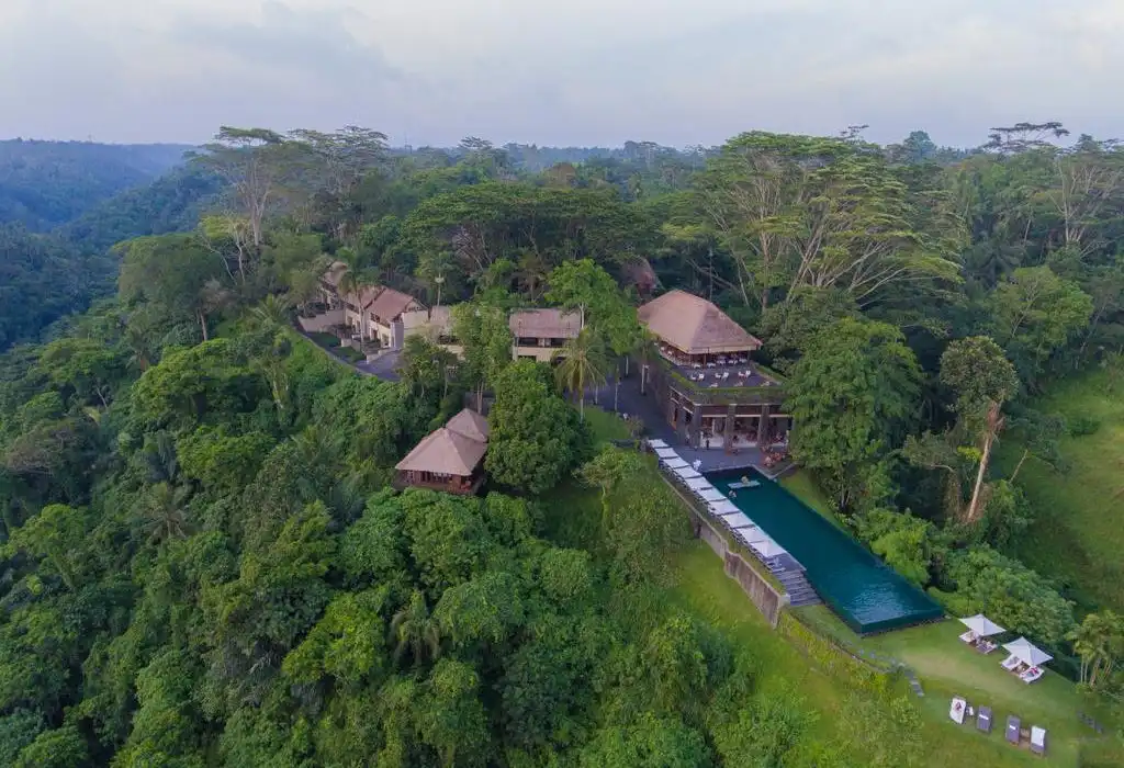 A drone photograph of a resort perched on the edge of a deep tropical rainforest valley. The property features traditional thatched-roof buildings, terraced lawns, and a striking, dark-tiled infinity pool extending out over the hillside, showcasing the dramatic scenery typical of the best luxury resorts in Bali.