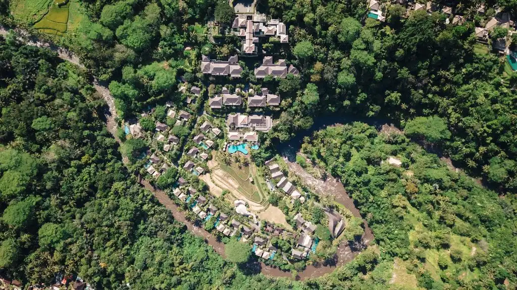 A high-altitude aerial view of an expansive resort complex nestled within a winding river bend and surrounded by dense tropical rainforest, featuring numerous private pool villas and terraced landscapes that showcase the stunning, secluded layouts typical of the best luxury resorts in Bali.