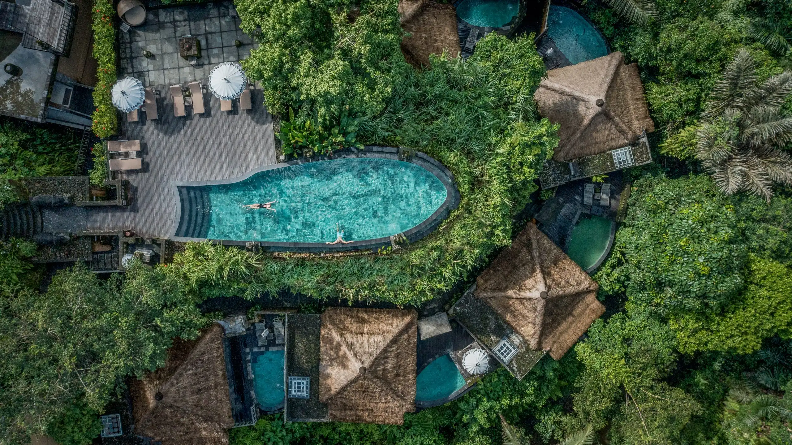 A top-down aerial view of a uniquely shaped infinity pool jutting out into a lush green jungle, surrounded by traditional thatched-roof villas and a wooden sun deck, showcasing the beautiful design of the best luxury resorts in Bali.