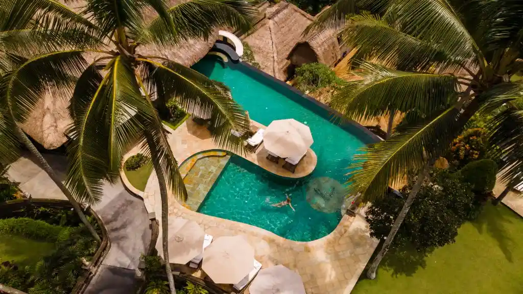 An aerial view of a person swimming in a vibrant turquoise, multi-level pool surrounded by tall palm trees, white sun umbrellas, and traditional thatched-roof villas, showcasing the stunning tropical amenities at resorts in bali.