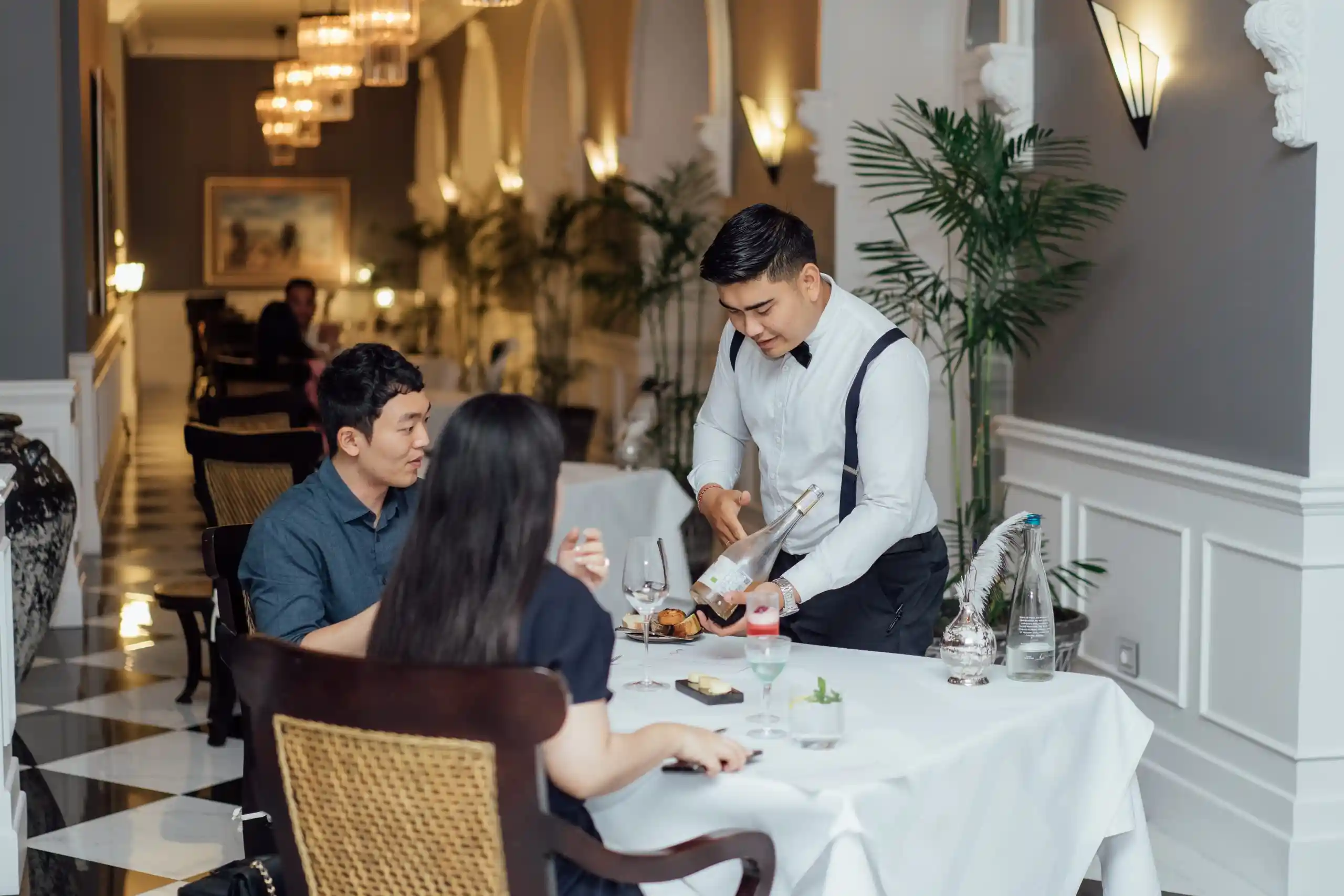 A waiter in a white shirt and black suspenders presents a bottle of wine to a couple seated at a white-clothed table in an elegant, warmly lit restaurant, highlighting the fine dining experiences available at luxury resorts in bali.