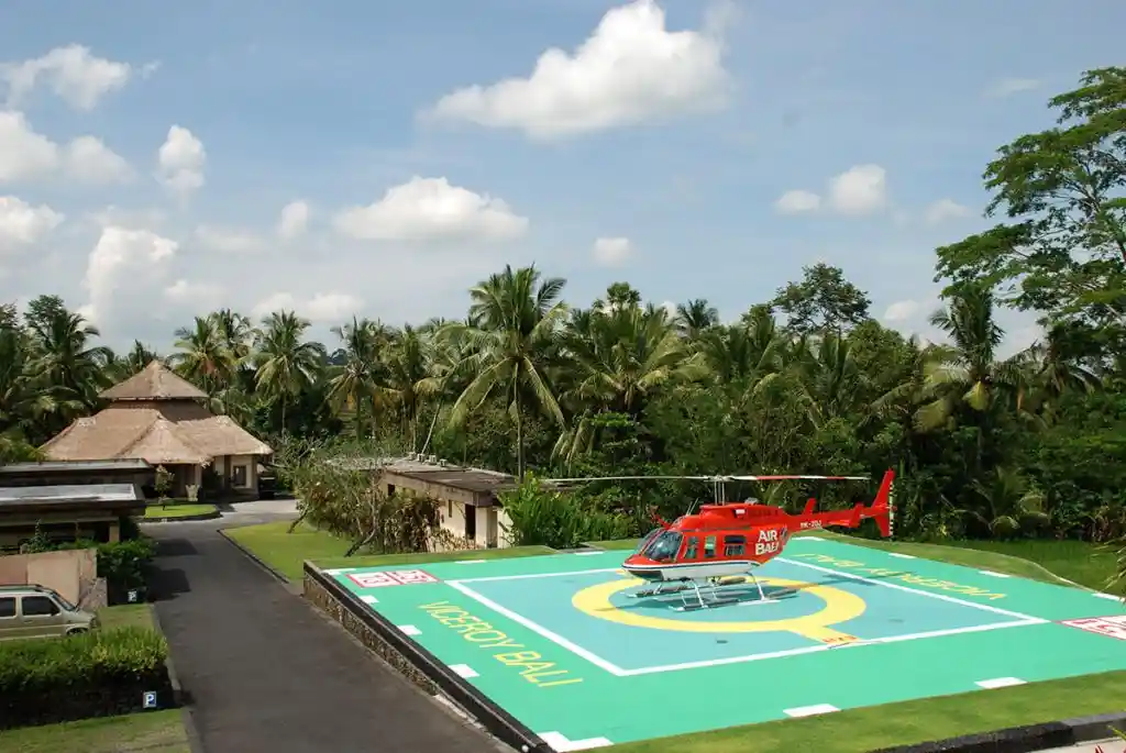 A bright red helicopter parked on a green landing pad labeled "Viceroy Bali," set against a backdrop of lush palm trees and traditional thatched-roof resort structures under a partly cloudy sky, highlighting the exclusive VIP transfer options available at luxury resorts in bali.