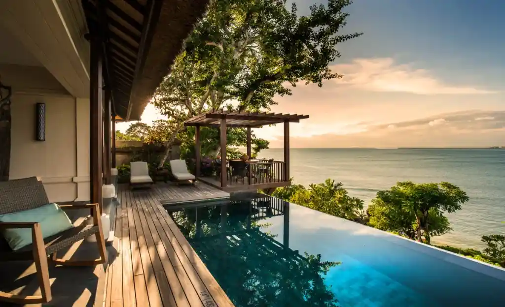 A luxurious private villa terrace featuring a wooden deck with lounge chairs, a dining pergola, and a stunning infinity pool overlooking a calm ocean under a soft golden sunset sky, showcasing the idyllic escapes offered by premium resorts in bali.