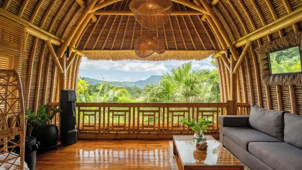 An exquisite open-air living room inside a bamboo villa featuring polished wooden floors, a modern grey sofa, woven hanging lamps, and a wide balcony offering panoramic views of lush tropical forests and distant mountains, showcasing the unique eco-luxury architecture of resorts in bali.