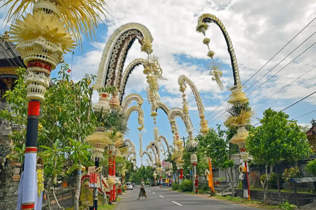 A vibrant street in Bali lined with numerous tall, intricately decorated, curving bamboo poles known as penjor. The poles are adorned with woven yellow and green palm leaves, arching gracefully over the road under a partly cloudy blue sky.