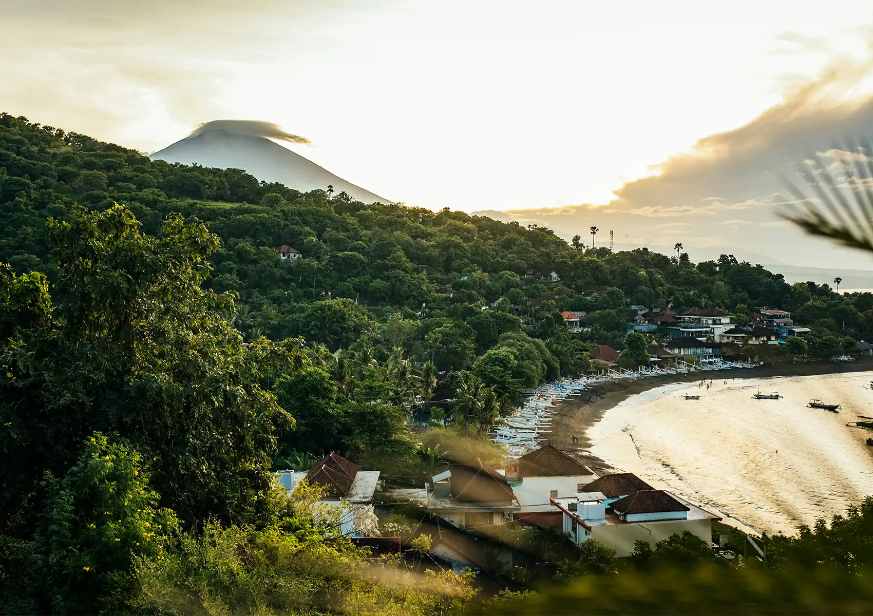 An elevated panoramic view of Amed village on Bali's east coast at golden hour, with rows of traditional outrigger fishing boats lining a black sand beach, lush jungle-covered hillside, and the majestic cone of Mount Agung capped by a lenticular cloud in the background