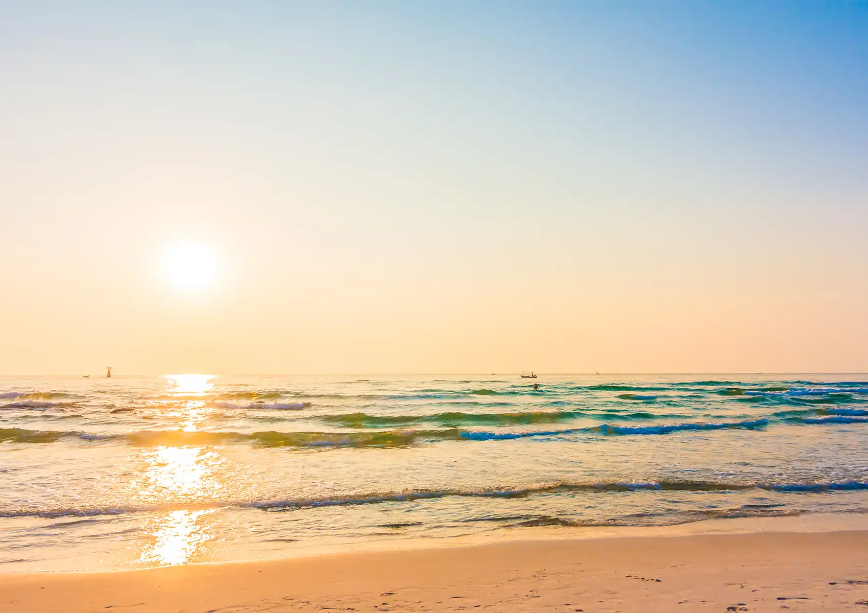 A tranquil tropical beach at sunrise with gentle turquoise waves lapping golden sand, the low sun casting a warm golden glow across the water, with a small traditional fishing boat visible on the horizon