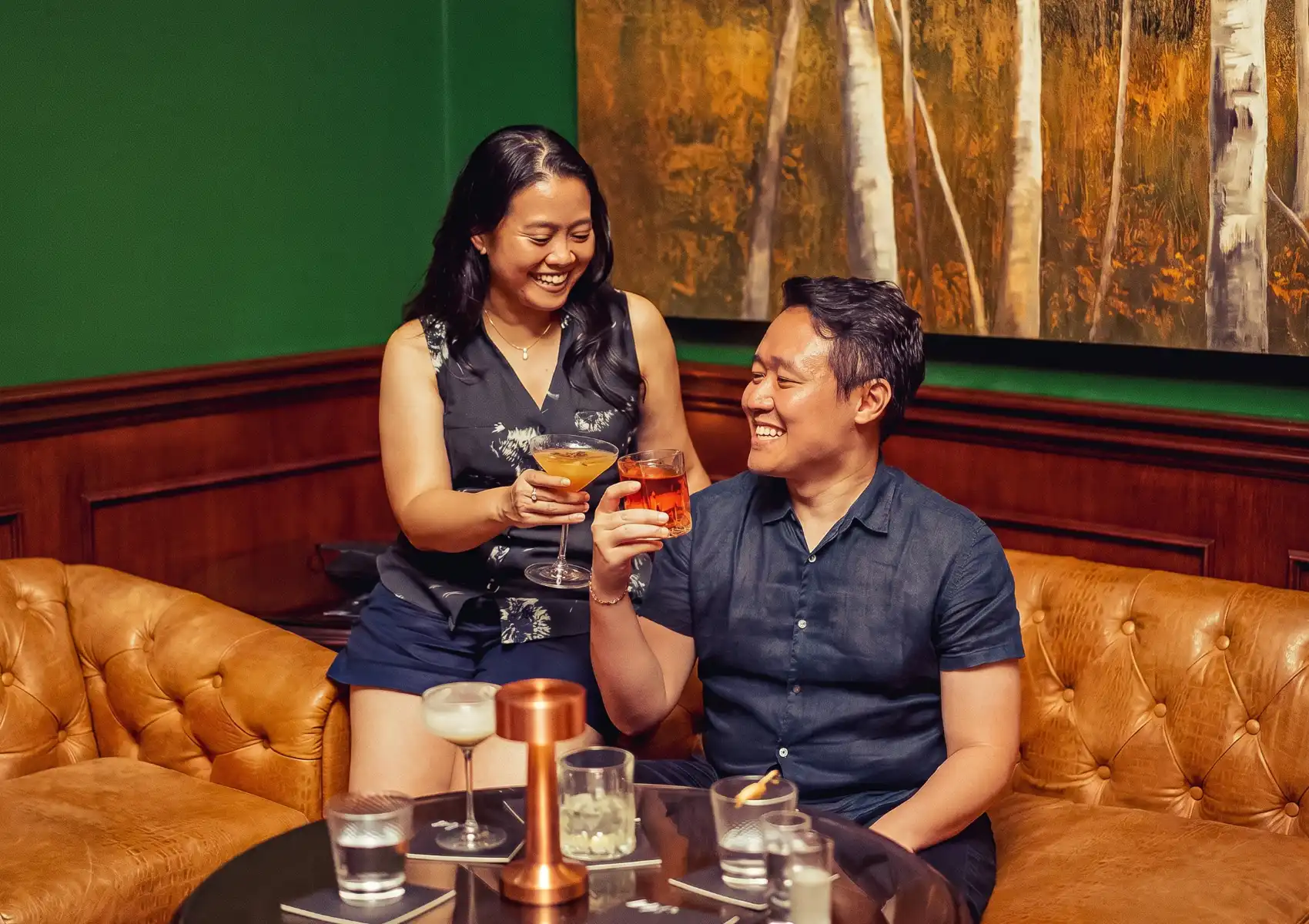 A joyful couple toasting with craft cocktails — an amber coupe and an orange negroni — on a tufted caramel leather sofa inside a moody bar with dark green walls, wood panelling, and an autumn forest oil painting, with additional drinks and a copper candle holder on the table