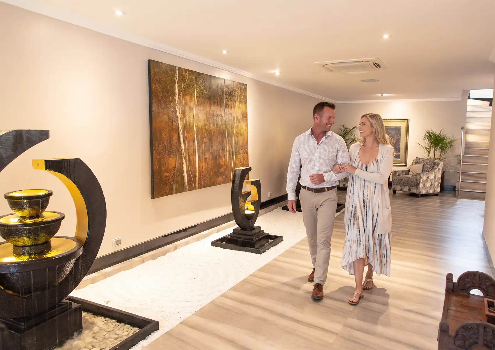 A smiling couple walking arm-in-arm through a modern boutique hotel lobby adorned with sculptural black-and-gold water features, white pebble garden beds, an autumn forest oil painting, and warm timber flooring leading toward a lounge area and staircase