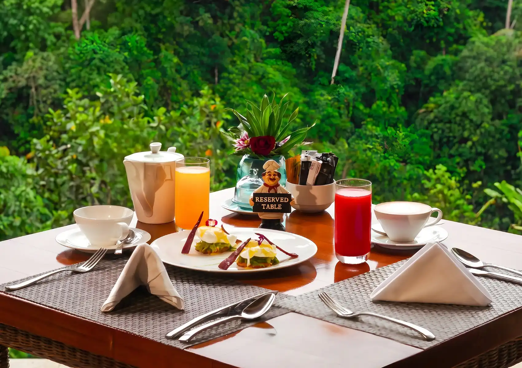 A neatly set outdoor breakfast table for two overlooking a dense tropical rainforest, featuring eggs Benedict with hollandaise sauce, fresh orange and red juices, a coffee cup, a teapot, folded white napkins, polished silverware, a tropical flower centrepiece, and a charming chef figurine holding a "Reserved Table" sign