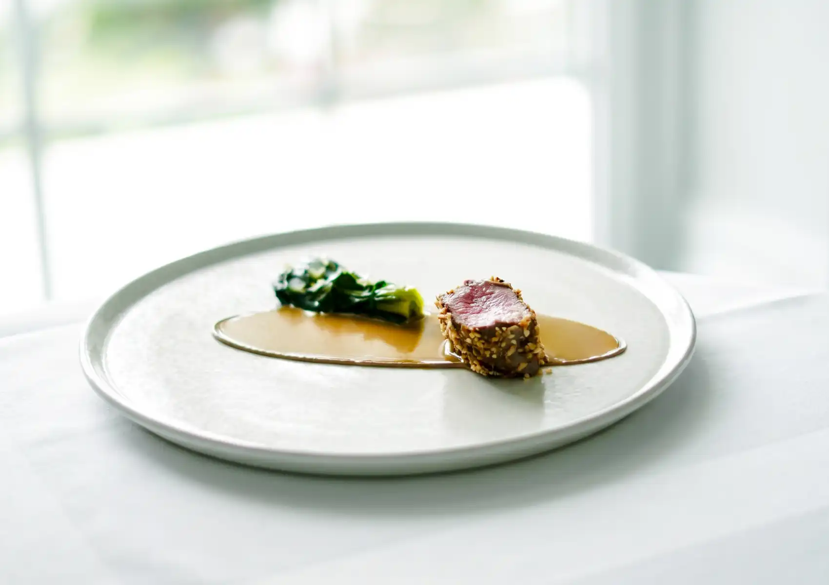 A minimalist fine dining plate on a white ceramic dish featuring a sesame-crusted medallion of rare seared meat sliced to reveal a pink centre, served alongside wilted greens in a pool of golden consommé, set against a softly lit white tablecloth with blurred greenery in the background