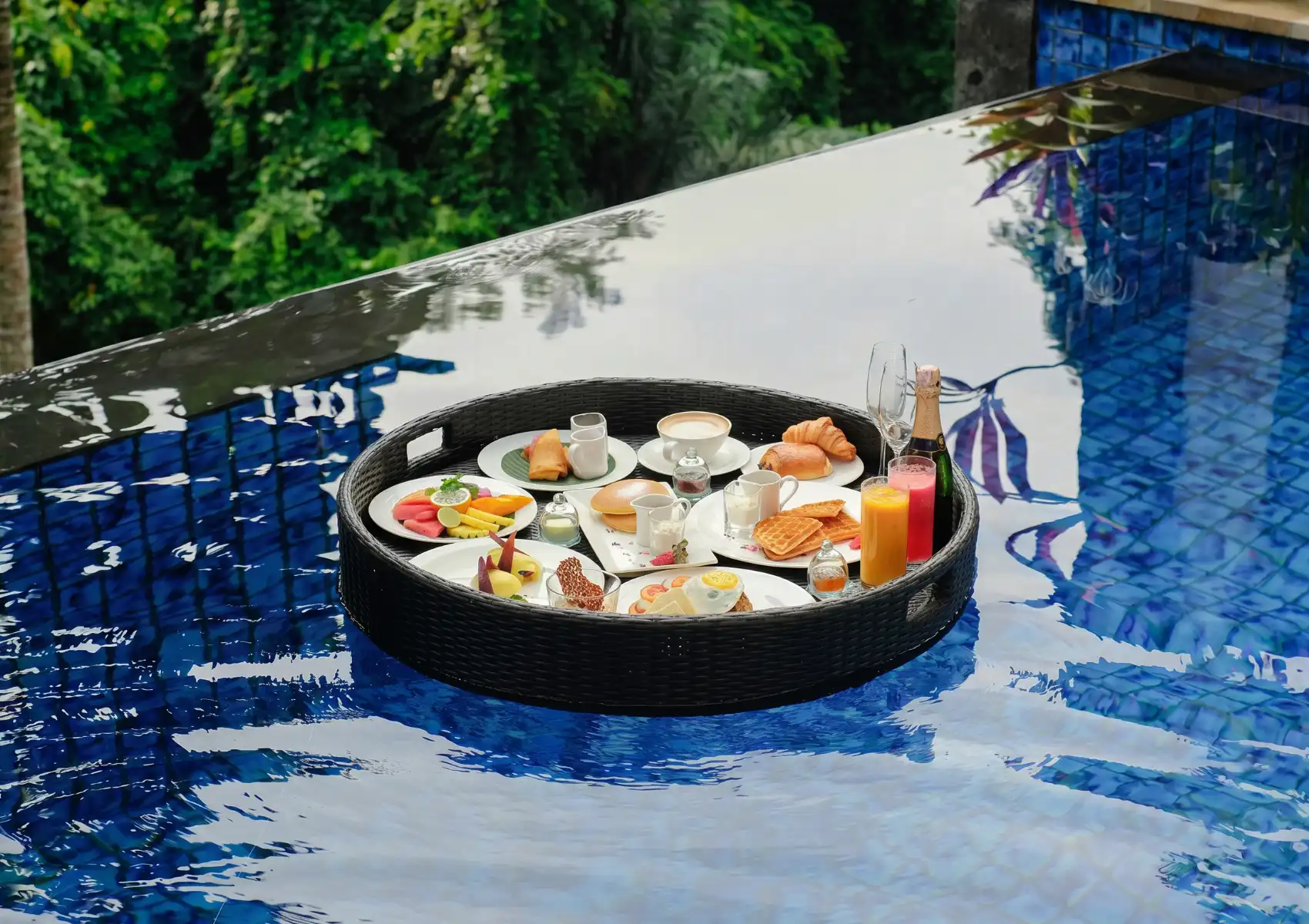 A breakfast spread of croissants, jam jars, and a cappuccino on an ornate wooden table inside a draped white-curtained cabana, overlooking a turquoise resort infinity pool where a woman stands at the edge gazing out over a misty jungle valley with traditional Balinese thatched-roof pavilions in the background