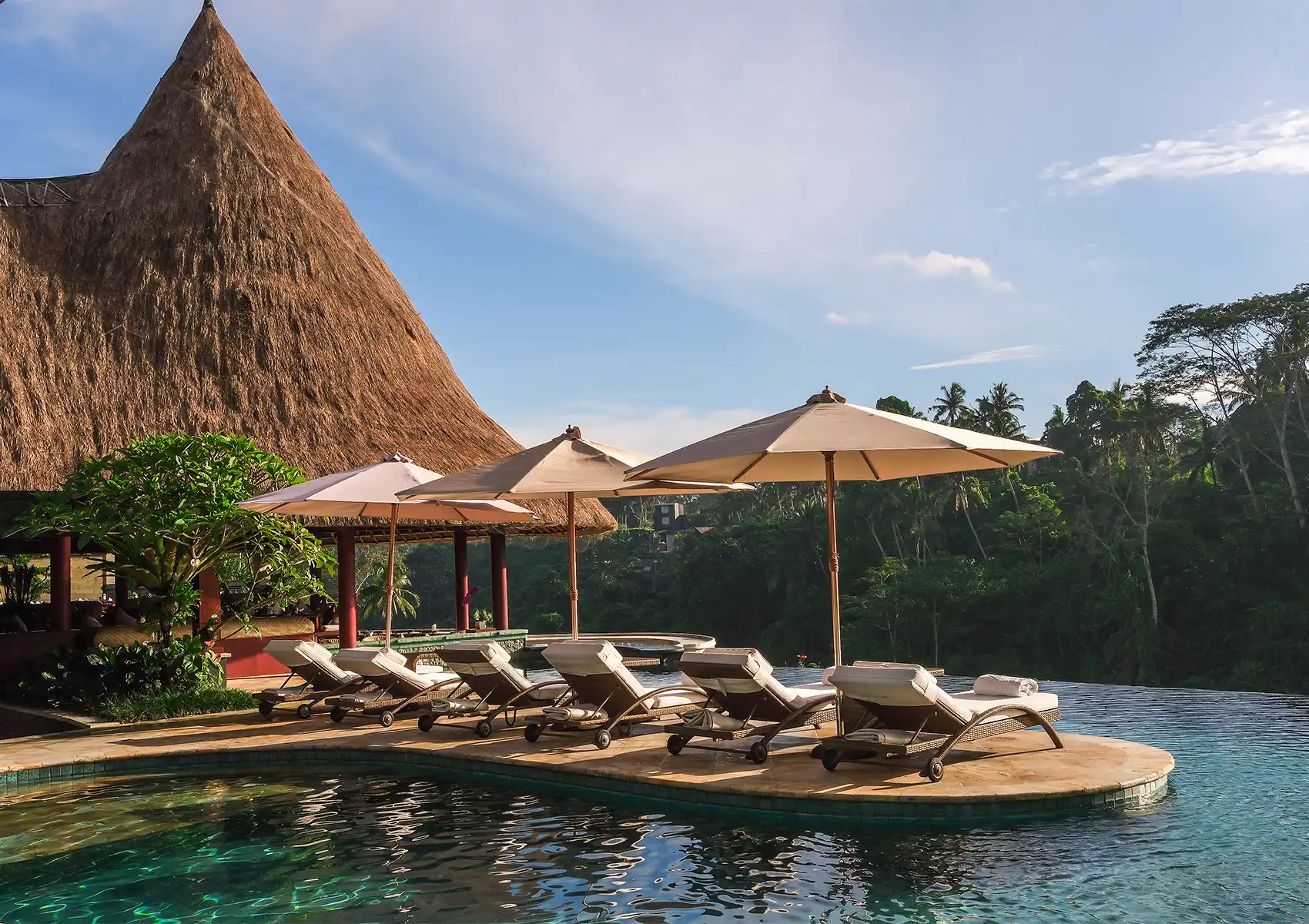A serene resort infinity pool edged with cushioned rattan sun loungers and beige parasols, flanked by a dramatic steep thatched roof pavilion and overlooking a lush tropical jungle valley under a partly cloudy blue sky