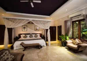 A spacious luxury villa bedroom featuring a king-size four-poster bed draped in sheer white canopy curtains, dark wood furniture, a ceiling fan over a deep purple tray ceiling, ornate chaise lounges, and a floor-to-ceiling window overlooking a lush stone-walled tropical garden