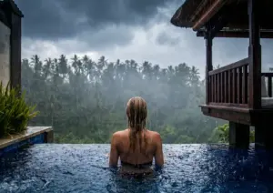 A woman seen from behind soaking in a private infinity pool during a heavy tropical rainstorm, with rain-drenched hair and rippling water in the foreground, a misty jungle of coconut palms in the distance, dramatic storm clouds overhead, and a traditional Balinese thatched gazebo to the right