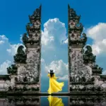A woman in a flowing yellow dress and wide-brimmed hat standing between the towering split gate of Pura Lempuyang temple, her reflection visible in the water below, with dramatic white cumulus clouds billowing against a vivid blue sky