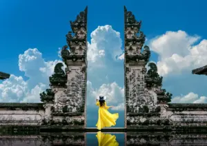 A woman in a flowing yellow dress and wide-brimmed hat standing between the towering split gate of Pura Lempuyang temple, her reflection visible in the water below, with dramatic white cumulus clouds billowing against a vivid blue sky