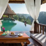 A breakfast spread of croissants, jam jars, and a cappuccino on an ornate wooden table inside a draped white-curtained cabana, overlooking a turquoise resort infinity pool where a woman stands at the edge gazing out over a misty jungle valley with traditional Balinese thatched-roof pavilions in the background