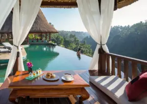 A breakfast spread of croissants, jam jars, and a cappuccino on an ornate wooden table inside a draped white-curtained cabana, overlooking a turquoise resort infinity pool where a woman stands at the edge gazing out over a misty jungle valley with traditional Balinese thatched-roof pavilions in the background