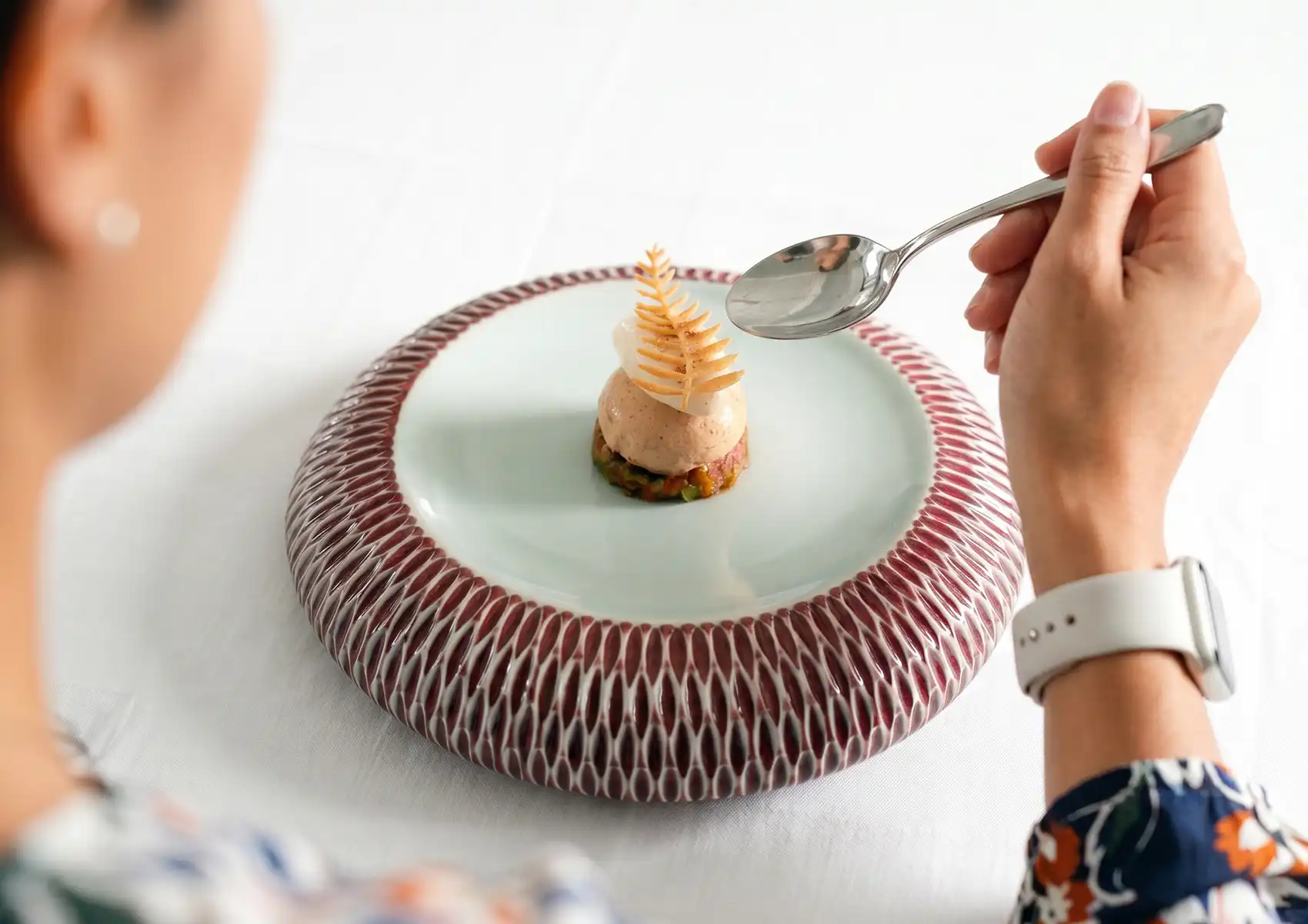 An over-the-shoulder shot of a person using a spoon to enjoy a beautifully plated, fine-dining gourmet dish topped with a delicate edible leaf garnish, highlighting the incredible culinary experiences available near your private pool villa Ubud.