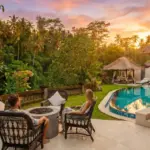 A couple relaxing in wicker chairs around a fire pit beside a private curved pool, enjoying a colorful jungle sunset at a boutique hotel Ubud.