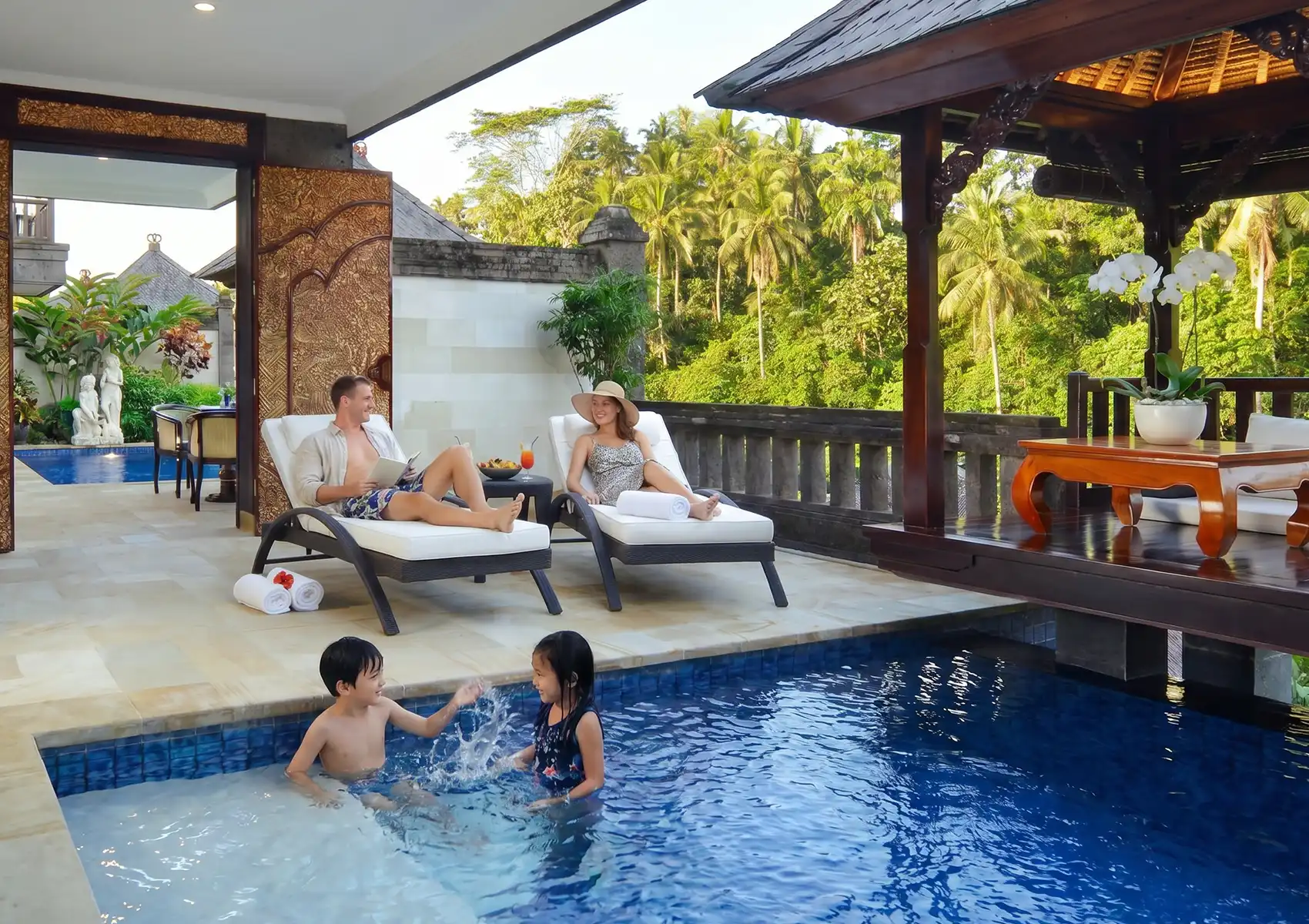 Parents relaxing on white lounge chairs beside a luxurious private villa pool while their two young children playfully splash in the water, capturing a tranquil moment on a family holiday in Ubud.