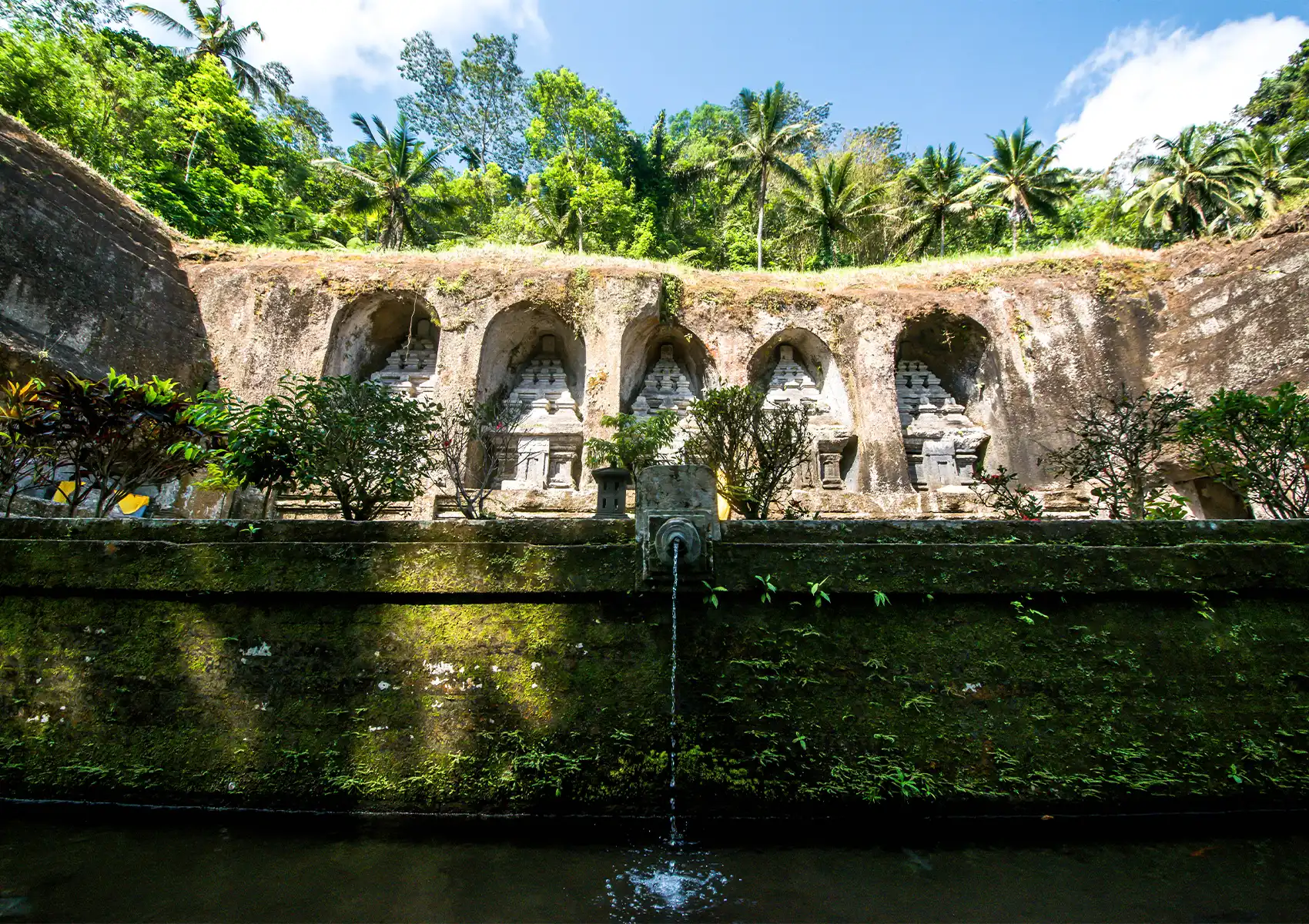 The ancient, majestic rock-cut shrines of Mount Kawi carved directly into a steep cliffside surrounded by lush tropical palm trees and a moss-covered water feature, a stunning historical landmark to explore while staying at a private pool villa Ubud.