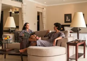A smiling couple in comfortable grey robes relaxing on a curved sofa in a spacious, luxurious hotel suite, enjoying a quiet morning during their 7 day bali honeymoon.