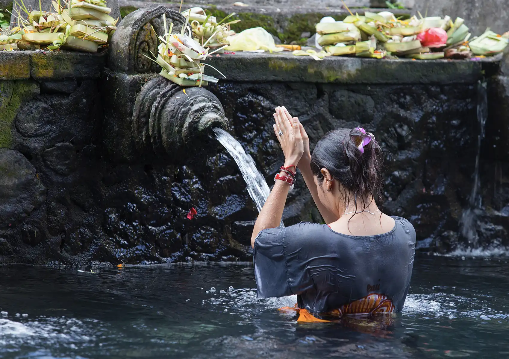 A woman standing waist-deep in a holy spring with her hands clasped in prayer, participating in a traditional Balinese water purification ritual under a carved stone spout, a profound cultural experience easily accessible from your private pool villa Ubud.