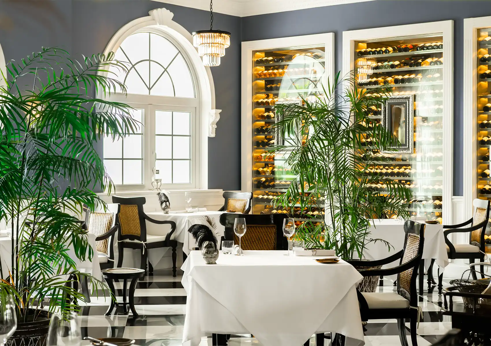 An elegant fine dining restaurant featuring black and white checkered floors, crisp white tablecloths, tropical indoor plants, and a massive illuminated wine cellar, offering a romantic dinner setting for a 7 day bali honeymoon.