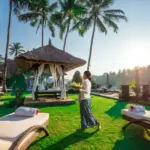 A woman in traditional Balinese attire carrying a tray walks across a lush, sunlit green lawn past white lounge chairs and a thatched-roof gazebo, highlighting the beautiful resort environment associated with sustainable travel Bali.