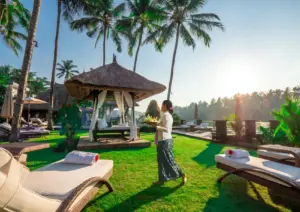 A woman in traditional Balinese attire carrying a tray walks across a lush, sunlit green lawn past white lounge chairs and a thatched-roof gazebo, highlighting the beautiful resort environment associated with sustainable travel Bali.