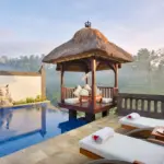 A breathtaking morning view from a luxury private pool villa Ubud, featuring a shimmering infinity pool, a traditional thatched-roof gazebo, and white lounge chairs with refreshing cocktails overlooking a misty, lush green jungle valley.