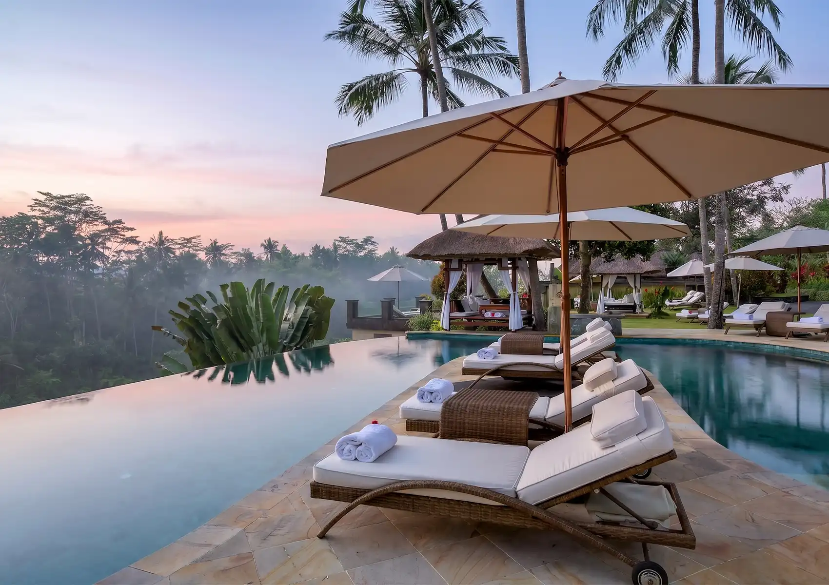 A tranquil resort infinity pool lined with lounge chairs and umbrellas overlooking a misty jungle valley at dawn, ideal for a relaxing stay on a bali trip itinerary 10 days.