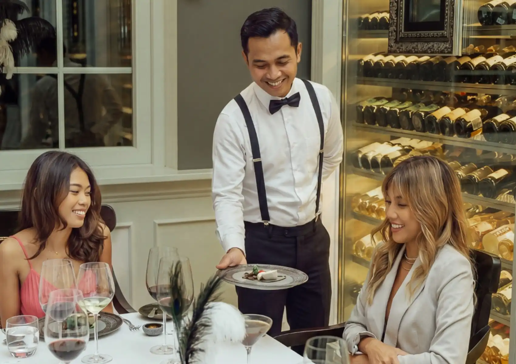 A smiling waiter in a white shirt and suspenders serving a gourmet dish to two women enjoying an elegant dinner during their luxury stay at a Private pool villa Ubud.