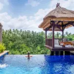 A woman relaxing in the infinity pool of a luxury private pool villa Ubud, looking out over a lush green jungle valley next to a traditional thatched-roof gazebo.