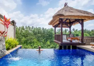 A woman relaxing in the infinity pool of a luxury private pool villa Ubud, looking out over a lush green jungle valley next to a traditional thatched-roof gazebo.