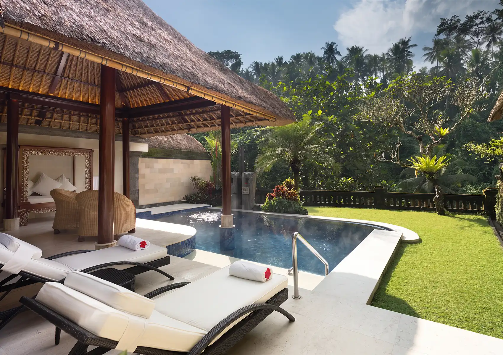 White lounge chairs and a traditional thatched-roof pavilion beside a steaming heated swimming pool at a tropical private pool villa Ubud surrounded by lush green landscaping.