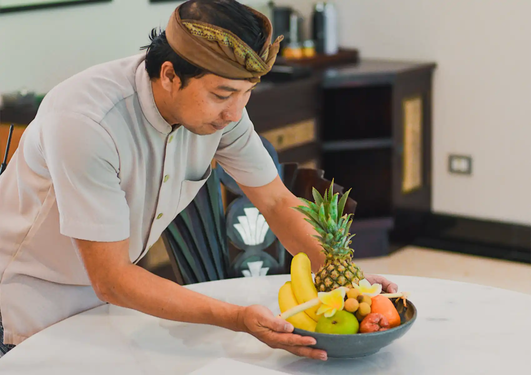 A welcoming staff member in traditional Balinese attire carefully placing a fresh tropical fruit bowl on a table at a luxury retreat Ubud.