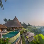 A stunning infinity pool and traditional thatched-roof villas perched on a hillside overlooking a misty, sunlit tropical valley at a breathtaking luxury retreat Ubud.
