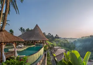 A stunning infinity pool and traditional thatched-roof villas perched on a hillside overlooking a misty, sunlit tropical valley at a breathtaking luxury retreat Ubud.