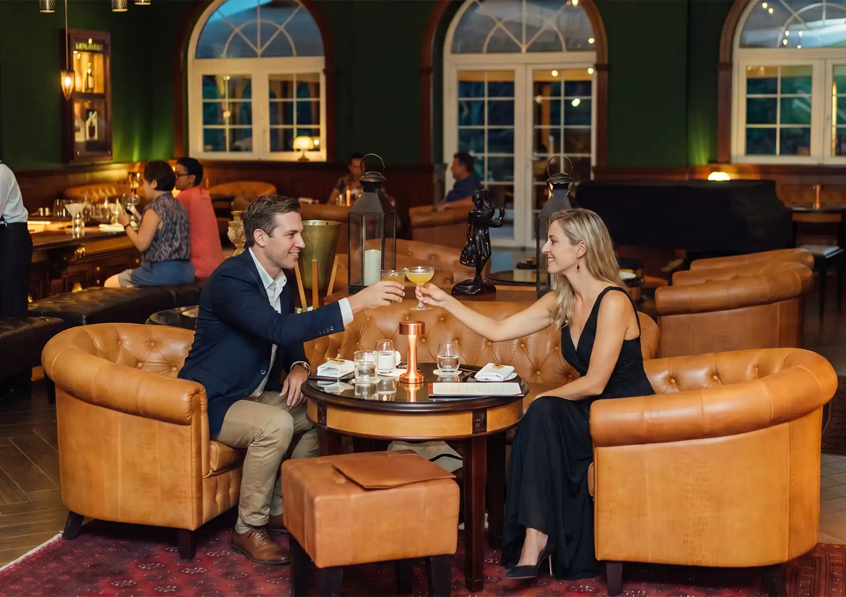 A couple dressed in formal attire toasting with cocktails in comfortable leather armchairs at an elegant lounge bar, showcasing evening things to do in Ubud.