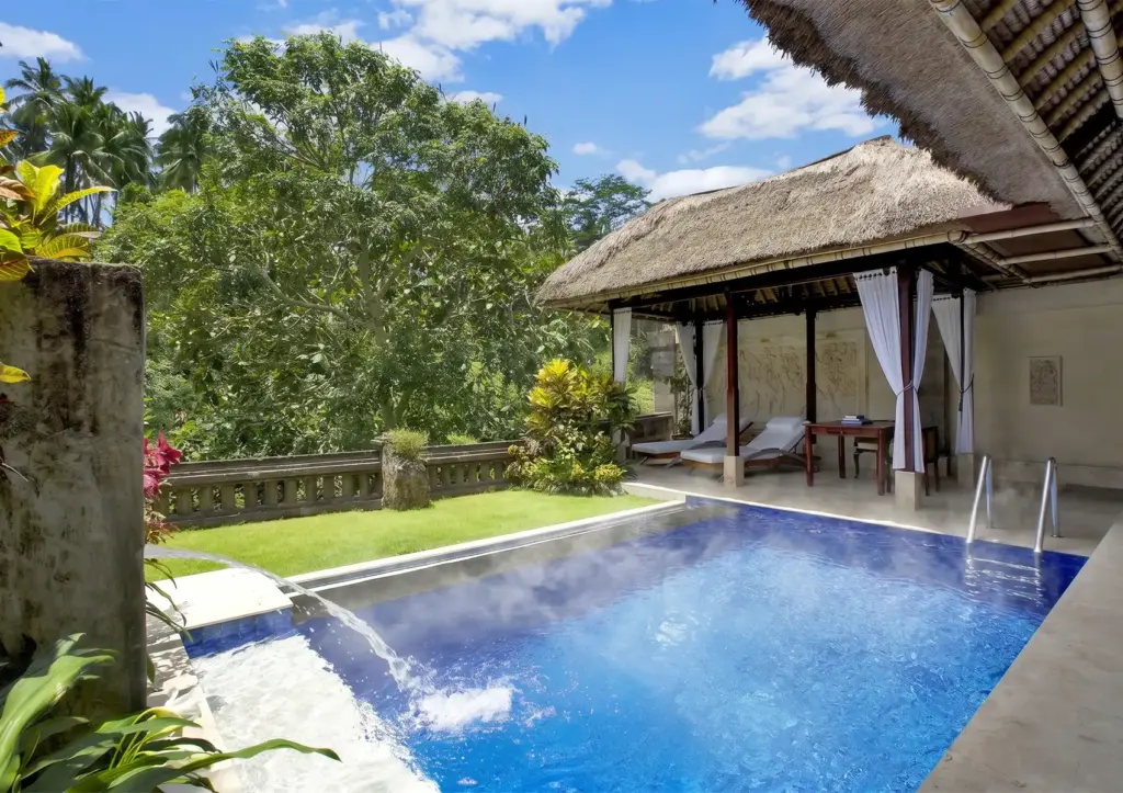 A steaming heated swimming pool with a flowing water feature alongside a relaxing thatched-roof gazebo surrounded by lush jungle foliage at a beautiful boutique hotel Ubud.