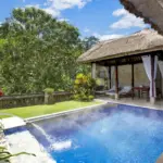 A steaming heated swimming pool with a flowing water feature alongside a relaxing thatched-roof gazebo surrounded by lush jungle foliage at a beautiful boutique hotel Ubud.